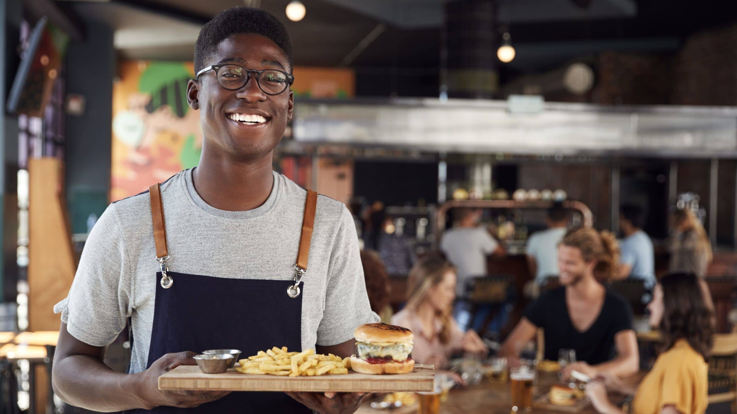 A smiling server carrying a tray with a burger and fries through the dining room